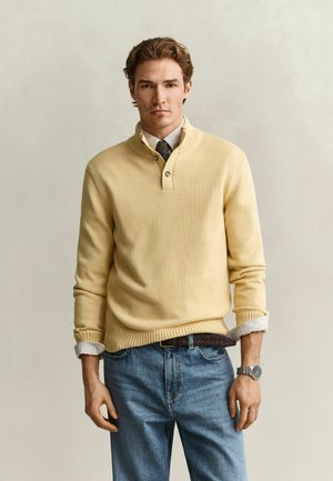 Homme debout, portant un pull jaune clair sur une chemise à col et une cravate, un jean bleu, une ceinture marron et une montre-bracelet argentée, fond neutre.