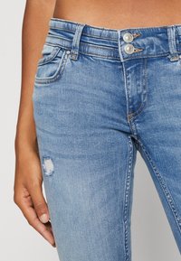 Jeans de mezclilla azul claro con corte ajustado, que cuentan con una cremallera de botones, dos bolsillos delanteros y un detalle desgastado en el muslo izquierdo.