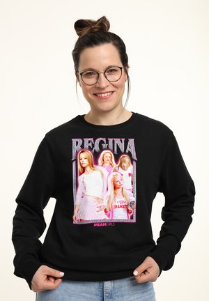 Kobieta z okularami, włosy związane w kok, uśmiechnięta, nosząca czarną bluzę z nadrukiem "Regina Mean Girls" przedstawiającym cztery blond kobiety w różowych strojach.