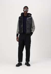 Vest negru cu umplutură și glugă peste un pulover din dungi bleumarin și alb, asortat cu pantaloni cargo largi negri și pantofi negri, accesorizat cu ochelari de soare.