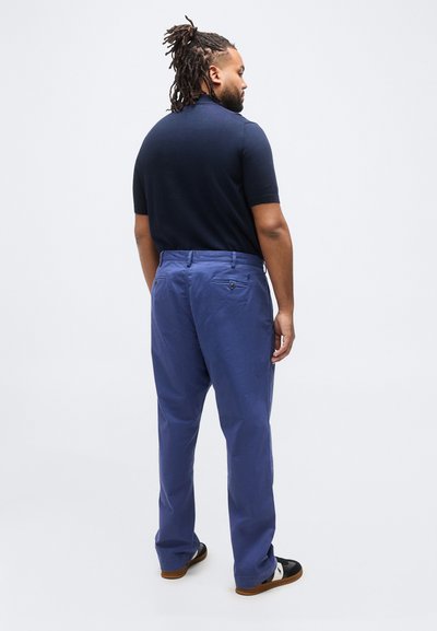 Άνδρας με κοτσίδες dreadlocks, ντυμένος με μπλε πόλο, μπλε παντελόνι και ασπρόμαυρα αθλητικά παπού τσια, στέκεται με την πλάτη προς τα έξω μπροστά σε λευκό φόντο.