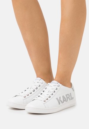 Zapatillas blancas de cuero con una textura lisa, que presentan una marca perforada en el lateral, puntera redonda y cordones blancos.