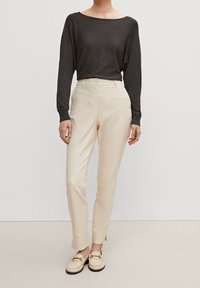 Pull noir à manches longues, pantalon crème fuselé avec plis sur le devant, et mocassins beige clair ornés de détails décoratifs en métal.
