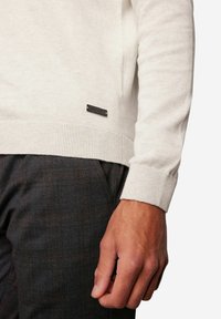 Hellgrauer Pullover aus weichem Stoff mit gerippten Bündchen und einem kleinen schwarzen Markenetikett nahe dem Saum. Kombiniert mit dunkelkarierter Hose.
