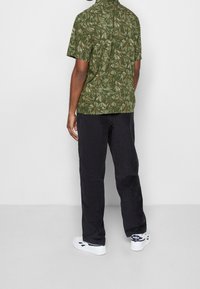 Camicia verde a maniche corte con motivo a foglie, abbinata a pantaloni neri larghi e sneakers bianche con dettagli blu.