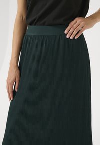 Gonna plissettata verde scuro con un ampio elastico in vita. Il tessuto presenta una texture a coste ed è progettata per cadere a metà polpaccio.