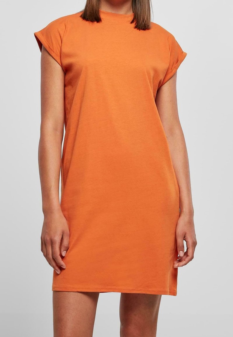 Une femme portant une robe orange décontractée à manches courtes, debout devant un fond gris clair uni.