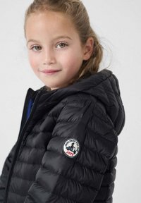 Giacca a vento nera con cappuccio, caratterizzata da una texture trapuntata, polsini a costine e una toppa con logo rotondo sul braccio superiore.