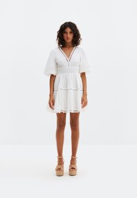 Moteris stovi, dėvinti trumpą baltą suknelę su pūstomis rankovėmis ir smėlio spalvos pakulnes, priešais vientisą baltą foną.