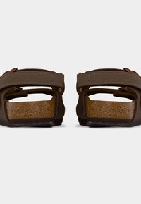 Καφέ σανδάλια με ρυθμιζόμενους ιμάντες και πατάκια από φελλό, εμφανιζόμενα από πίσω σε λευκό φόντο.