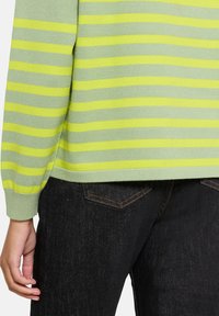 Jasnozielony sweter w paski z żółtymi poziomymi pasami, ściągaczami przy rękawach i lekko skróconym dołem, zestawiony z czarnymi jeansami denim.