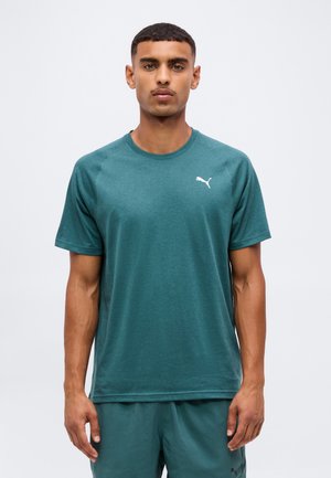 Homme portant un maillot de sport turquoise Puma à manches courtes et un short assorti, debout devant un fond blanc uni.