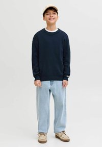 Garçon souriant portant une casquette beige, un sweat-shirt bleu marine, un jean large bleu clair et des chaussures beige à lacets, debout devant un fond clair uni.