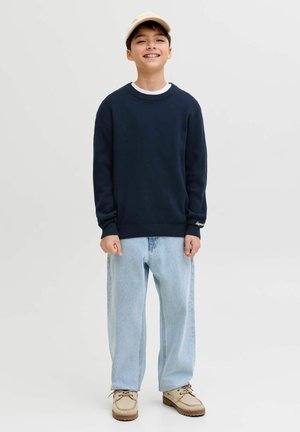 Usmívající se chlapec v béžové čepici, námořnicky modré mikině, světle modrých širokých džínách a béžových šněrovacích botách stojí na jednoduchém světle pozadí.