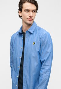 Μπλε και λευκό ριγέ μακρυμάνικο πουκάμισο από βαμβάκι, με γιακά με κουμπιά και κεντημένο κίτρινο λογότυπο στο στήθος.