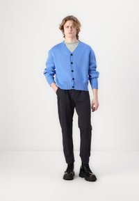 Cardigan tricotat albastru, cu guler în V, închidere cu nasturi și manșete cu nervuri. Purtați peste o cămașă gri deschis și asortat cu pantaloni negri și bocanci.