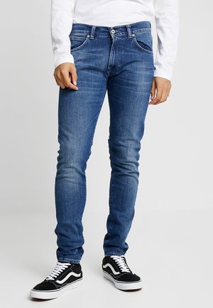 Mężczyzna noszący niebieskie dopasowane jeansy, białą koszulę z długim rękawem oraz czarne niskie trampki z białymi sznurówkami i podeszwami.