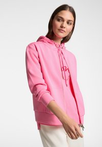 Felpa rosa con zip realizzata in tessuto morbido, dotata di cappuccio con coulisse, tasche frontali e un logo di colore contrastante sul petto.