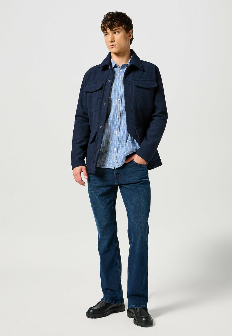 Giacca blu navy con due tasche sul petto, indossata sopra una camicia a quadri azzurra chiara. Abbinata a jeans di denim scuro e calzature nere.