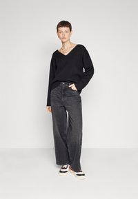 Crna v-neck pulover s visokim strukom, uparen s crnim trapericama širokih nogavica. Model nosi bijele tenisice s crnim i crvenim detaljima.
