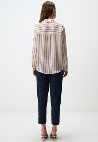 Camicia bianca a maniche lunghe con strisce verticali rosse, dotata di un colletto classico, piega posteriore e leggero arrotondamento dell'orlo. Abbinata a pantaloni navy.