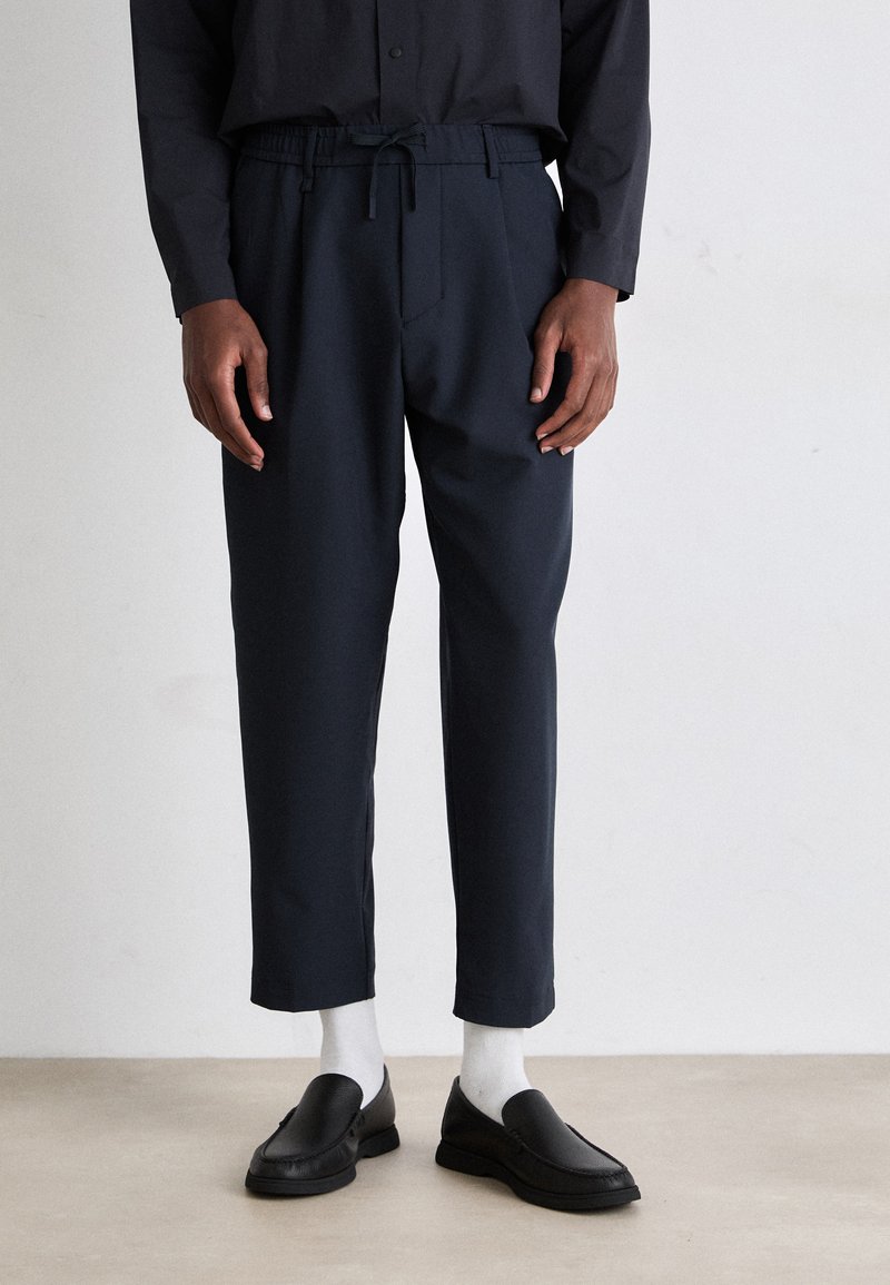 Tengerészkék, tapered szabású nadrág, rugalmas derékkal és zsinórral. Fekete slip-on cipőkkel és fehér zoknikkal párosítva. Sima anyag, minimalista dizájn.