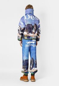 Chaqueta acolchada y pantalones a juego con un patrón de paisaje azul con texto, acentuados por un cuello alto y puños elásticos. Botas marrones completan el conjunto.