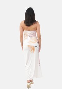 Femme aux cheveux raides et foncés portant une robe blanche sans bretelles avec un imprimé floral pêche et des sandales à talons blancs, se tenant de dos.