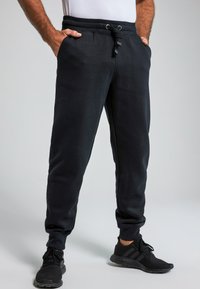 Pantalones de chándal negros con cintura elástica y cordón, con bolsillos laterales y piernas ajustadas. La tela parece suave y texturizada.