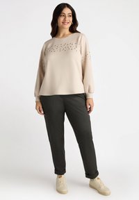Beige sweatshirt met decoratieve rhinestones op de buste en mouwen, gecombineerd met donkergrijze broek en crèmekleurige sneakers. Glad stofoppervlak.