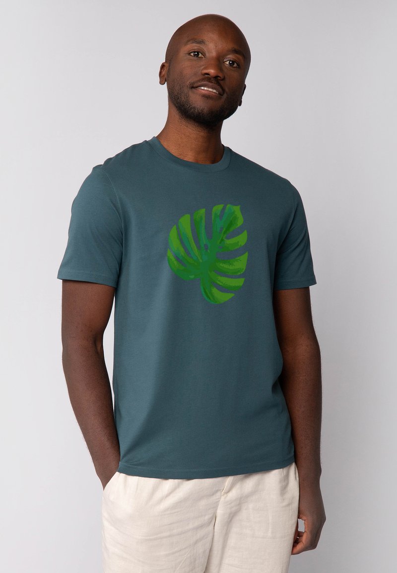 Teal kleurige t-shirt met korte mouwen gemaakt van zacht materiaal, met een grote groene monstera bladprint op de voorkant. Eenvoudig ronde hals ontwerp.