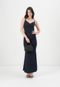 Vestido maxi justo azul marinho com alças finas tipo espaguete, combinado com uma mala preta texturizada e sandálias pretas com tiras.