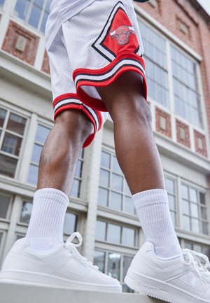 Osoba ubrana w białe spodenki do koszykówki Chicago Bulls, białe skarpetki i białe trampki stojąca na zewnątrz budynku z czerwonej cegły z dużymi oknami.