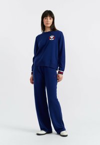 Marineblauwe gebreide sweatshirt met een ronde logo, gecombineerd met bijpassende wijde broek. Beschikt over rode en witte gestreepte manchetten. Zachte textuur.