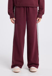 Burgundy sweatpants with a relaxed fit, featuring white side stripes, soft texture, and elastic waist. Worn with light-colored sneakers.
