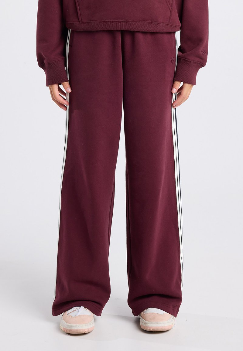 Burgundy sweatpants with a relaxed fit, featuring white side stripes, soft texture, and elastic waist. Worn with light-colored sneakers.