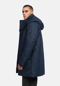 Tums zilas izolēta parka ar kapuci, priekšējiem kabatām un logo plāksteri uz piedurknes. Gluda tekstūra ar spīdīgu apdari.