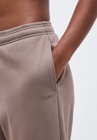 Το χέρι ενός ατόμου ακουμπάει μέσα στην τσέπη ενός τζόγκινγκ παντελονιού ταουπ με ελαστική μέση και το λογότυπο "BORG" στο ισχίο.