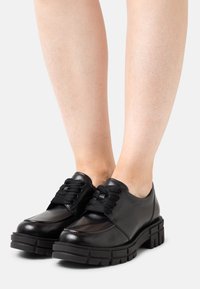 Chaussures en cuir à semelle épaisse noires avec des lacets épais, portées pieds nus, montrées de face et de côté.