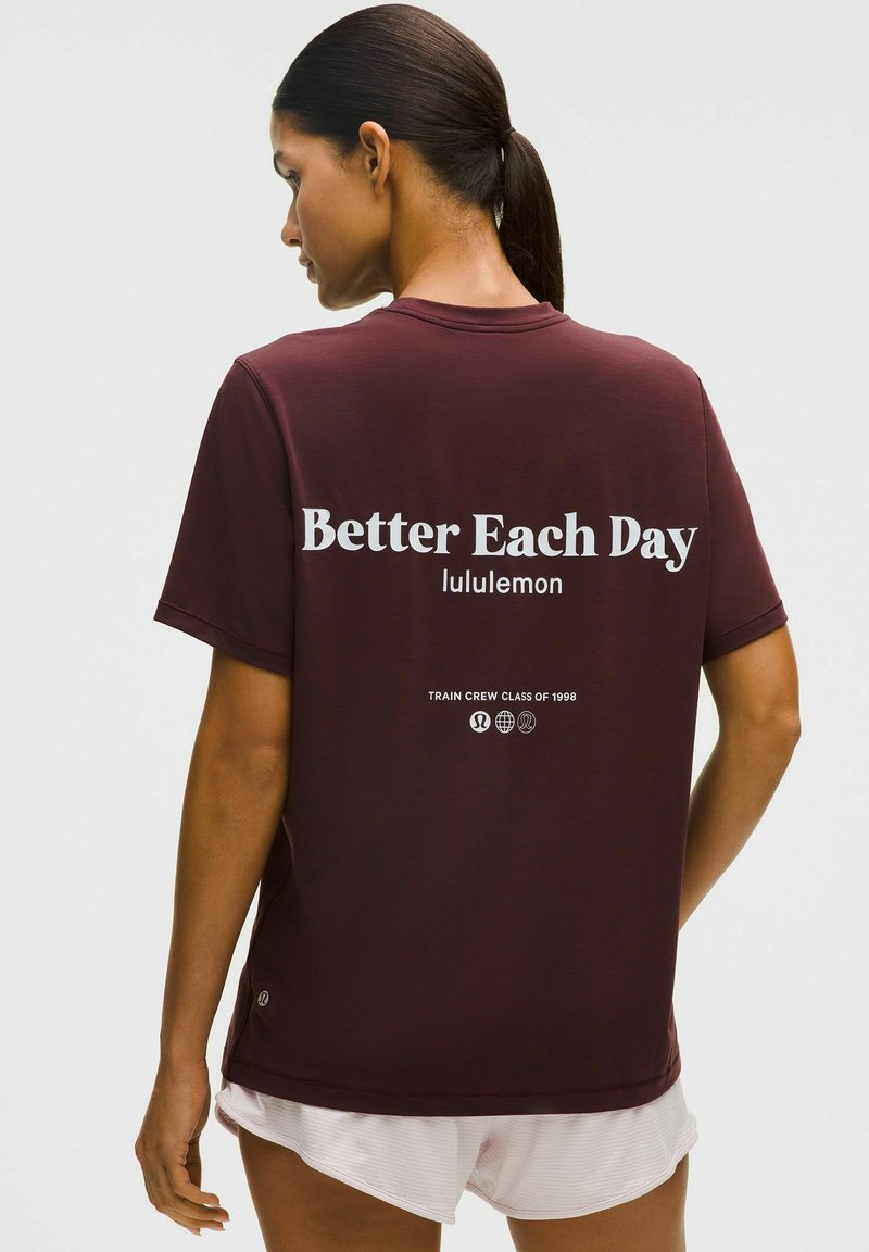 Sötét hajú nő lófarokkal, bordó "Better Each Day lululemon" pólóban és fehér rövidnadrágban, háttal áll egy sima háttér előtt.