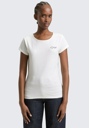 Kobieta z plecionymi włosami, ubrana w białą koszulkę z napisem "Smile" na piersi, w ciemnoniebieskich jeansach, stojąca na jednolitym tle.