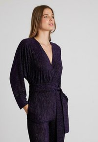 Conjunto estilo envolvente en color púrpura con mangas largas, que presenta una textura plisada y un cinturón a juego. La tela tiene un ligero brillo.
