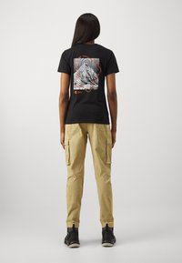 Svart bomullströja med grafiskt tryck på ryggen som har linjer och text i vitt och orange, kombinerad med beiga cargo-byxor.