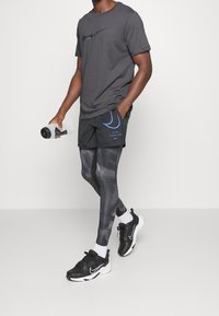 Czarny T-shirt Nike, czarne szorty z niebieskimi akcentami, wzorzyste czarne legginsy oraz czarne buty sportowe, trzymająca butelkę wody.