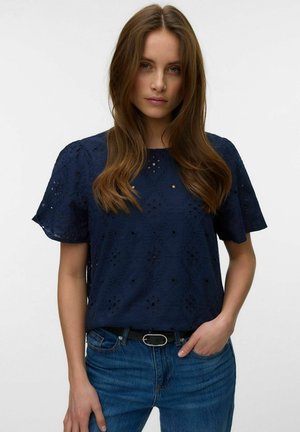 Jeune femme aux longs cheveux bruns portant un chemisier bleu marine à œillets et un jean bleu, debout avec une main dans la poche sur fond blanc.