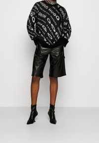 Zwart-grijs gebreide trui met patroon en tekst "HUGO", gecombineerd met hooggetailleerde zwarte leren shorts en zwarte puntige enkellaarsjes.