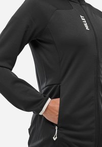 Veste noire zippée avec un tissu texturé, des poches latérales et un détail en blanc au niveau des poignets intérieurs. Comprend un logo discret sur le devant.
