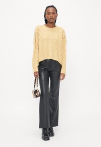 Pull en maille torsadée jaune clair avec des manches longues, associé à un pantalon flair en simili cuir noir et un petit sac à main à motifs.