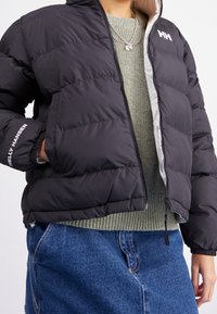 Personne portant une doudoune noire Helly Hansen par-dessus un pull en tricot vert et un pantalon en jean bleu, la main dans la poche de la veste.