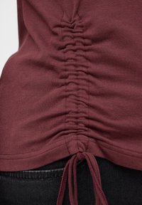 Bordeauxrotes Oberteil mit geraffter Textur am Rücken und verstellbaren Bändern, gefertigt aus weichem Stoff. Glatte Oberfläche und lässiges Design.